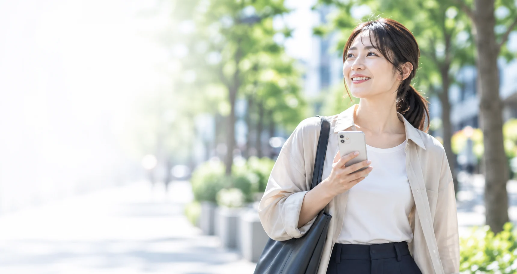緑のある街をスマートフォンを手に歩く女性